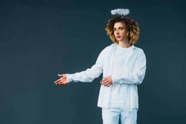 curly teenager in angel costume with halo above head pointing with hand isolated on green clipart