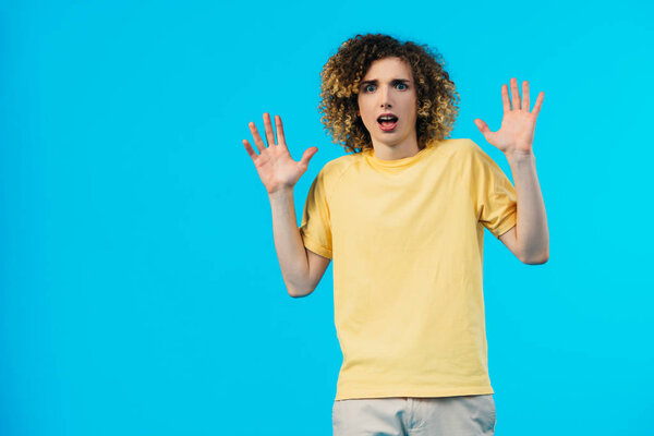 scared curly teenager showing hands isolated on blue