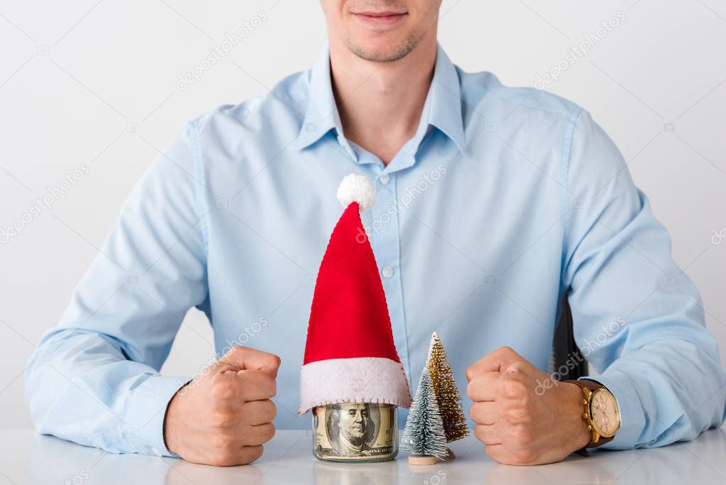 cropped view of man with clenched fists near glass jar with money and santa hat isolated on white 