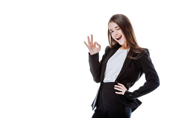 winking and smiling businesswoman in suit showing ok sign isolated on white