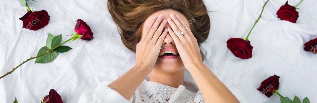 panoramic shot of smiling woman obscuring face and lying on bed 