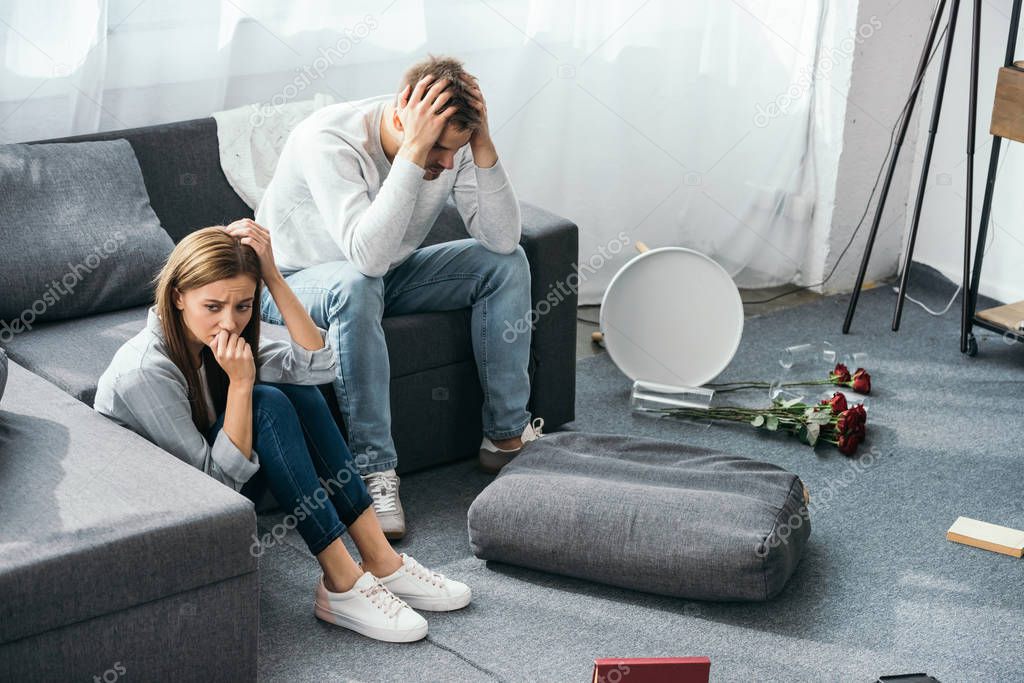 sad woman sitting on floor and handsome man sitting on sofa in robbed apartment 