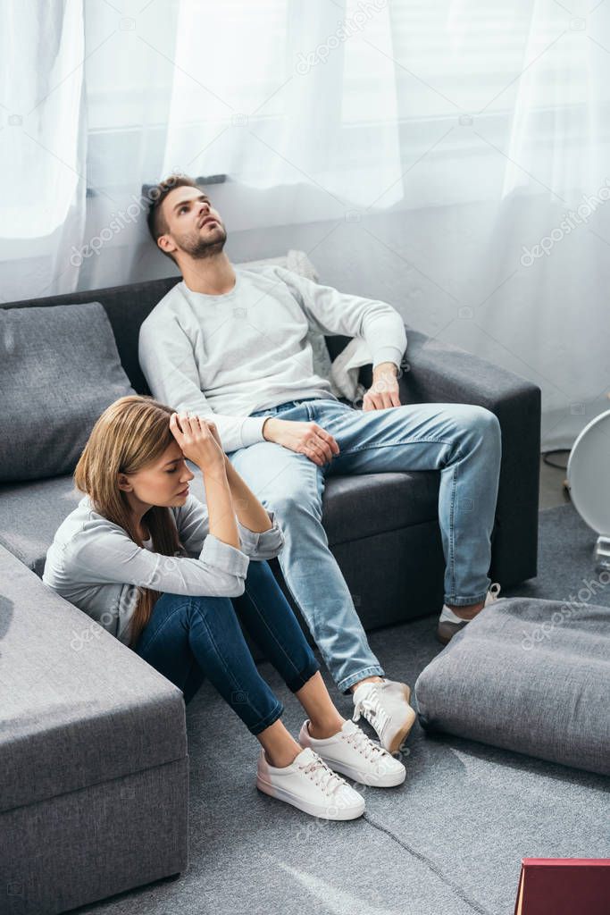 sad woman sitting on floor and handsome man sitting on sofa in robbed apartment 