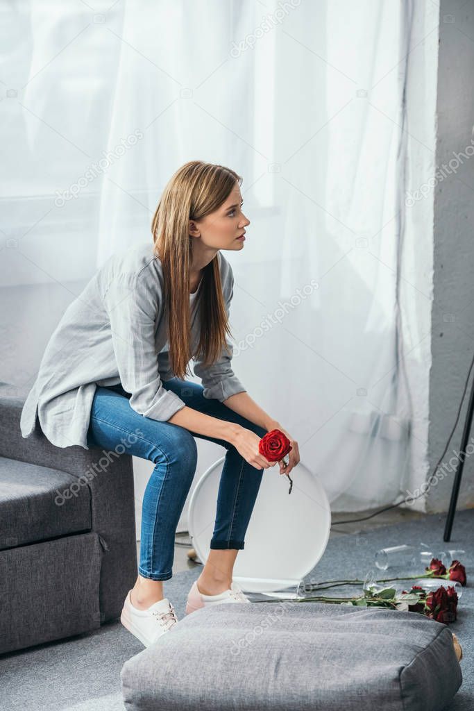 attractive woman sitting on sofa and holding rose in robbed apartment 