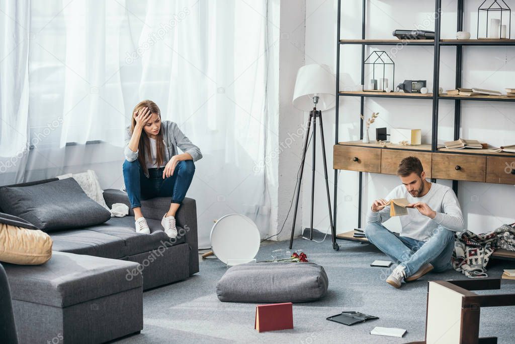 sad woman sitting on sofa and handsome man sitting on floor in robbed apartment 