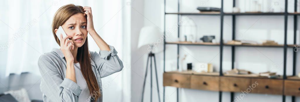 panoramic shot of attractive and sad woman talking on smartphone in robbed apartment 