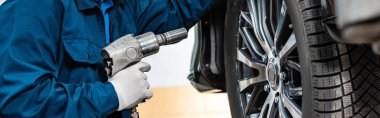 cropped view of mechanic holding pneumatic wrench near car wheel, panoramic shot clipart