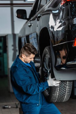Genç tamirci atölyede yükseltilmiş arabaya tekerlek takıyor.