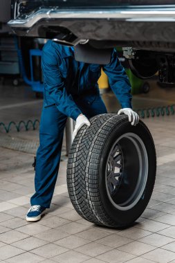 cropped view of mechanic with wheel near raised car in workshop clipart