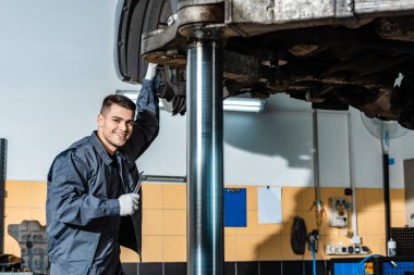 smiling mechanic holding wrench and looking at camera while standing under car raised on car lift clipart