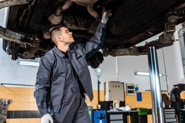 attentive mechanic inspecting bottom of raised car in workshop clipart
