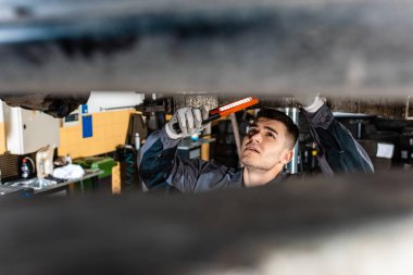 selective focus of mechanic inspecting car bottom with flashlight clipart