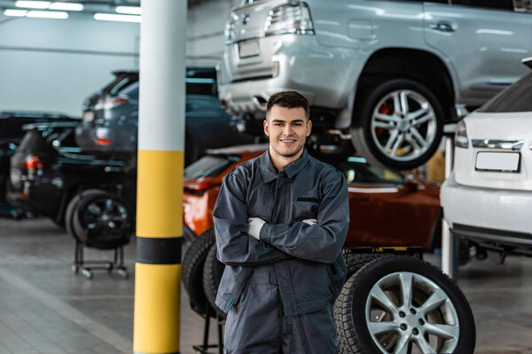 smiling mechanic looking at camera while standing in workshop with crossed arms