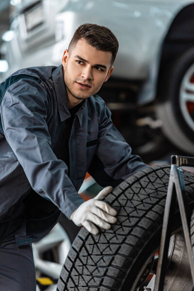 young confident mechanic looking away near car wheel in workshop