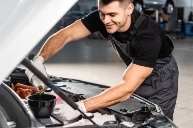 selective focus of smiling mechanic pouring oil to car engine clipart