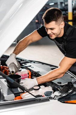 handsome, smiling mechanic pouring oil to car engine clipart