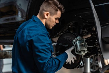 young, attentive mechanic installing disc brakes on raised car clipart