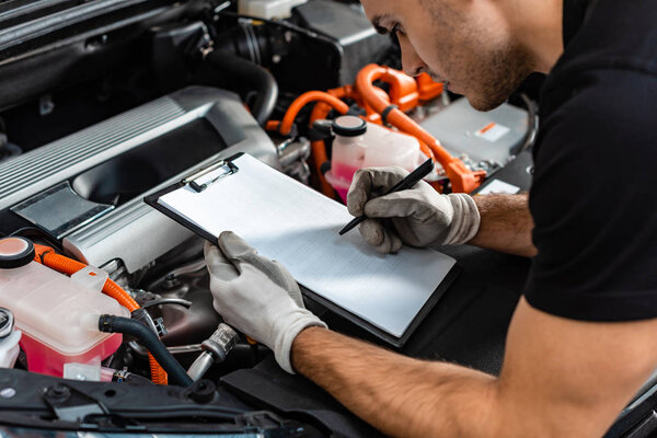 cropped view of mechanic writing on clipboard while inspecting car engine compartment