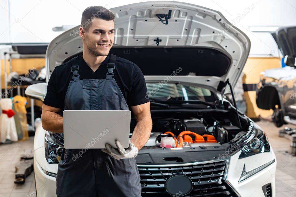 handsome mechanic holding laptop while standing near car with open hood