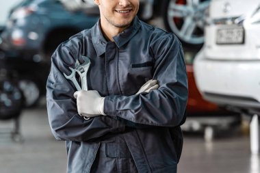 cropped view of smiling mechanic standing with crossed arms and holding wrenches clipart