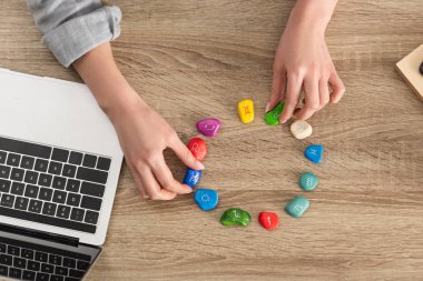 Cropped view of woman holding stones with zodiac signs beside laptop on table clipart