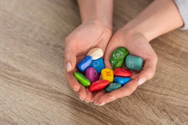 Cropped view of woman holding in hands colorful stones with zodiac signs at wooden table clipart