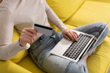 Kredi kartı tutan ve koltukta dizüstü bilgisayar kullanan bir kadın görüntüsü.