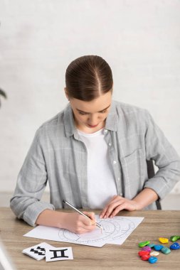 Astrologer drawing natal chart beside cards and stones with zodiac signs on table clipart