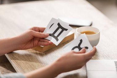 Kahvenin ve kitabın yanında zodyak tabelaları olan astrologların elindeki kartların kesilmiş görüntüsü.