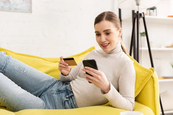 Chica Sonriente Usando Smartphone Sosteniendo Tarjeta Crédito Sofá Sala Estar —  Fotos de Stock