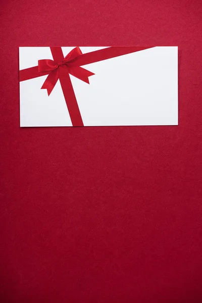コピースペース付きの赤い表面に弓付きの白いグリーティングカードのトップビュー — ストック写真