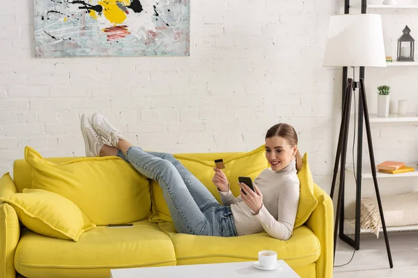 笑顔の若い女性がクレジットカードを保持し 自宅でソファにスマートフォンを使用して — ストック写真