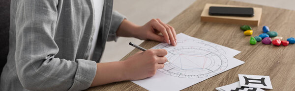 Cropped view of astrologer drawing natal chart by cards with zodiac signs on table, panoramic shot