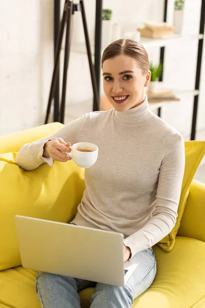 コーヒーを飲みながらカメラで微笑む魅力的な女性とソファの上でノートパソコンを使う — ストック写真