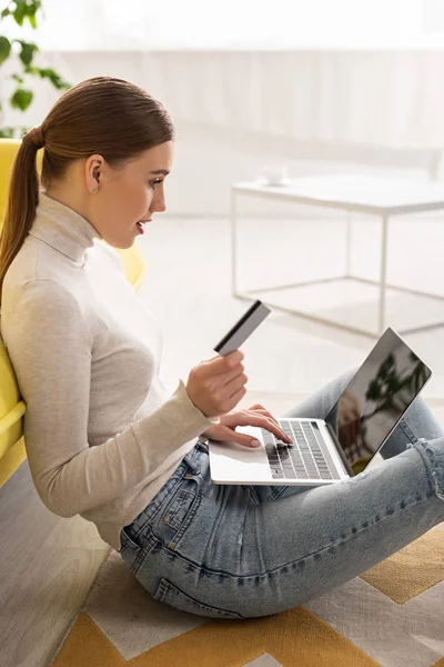 stock image Side view of beautiful woman holding credit card and using laptop on floor in living room