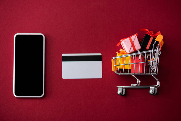 Top view of smartphone, credit card and toy gift boxes in cart on red background