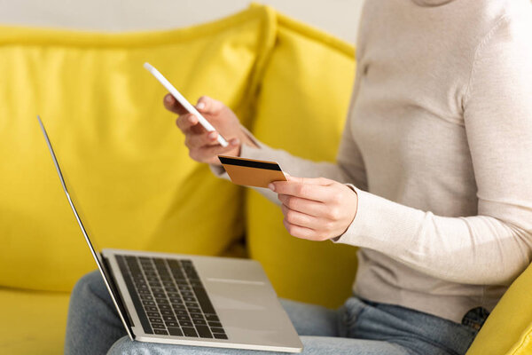 Cropped view of girl holding credit card, smartphone and laptop on sofa