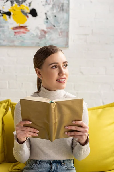 Sorrindo Mulher Segurando Livro Olhando Para Longe Sofá Sala Estar — Fotografia de Stock