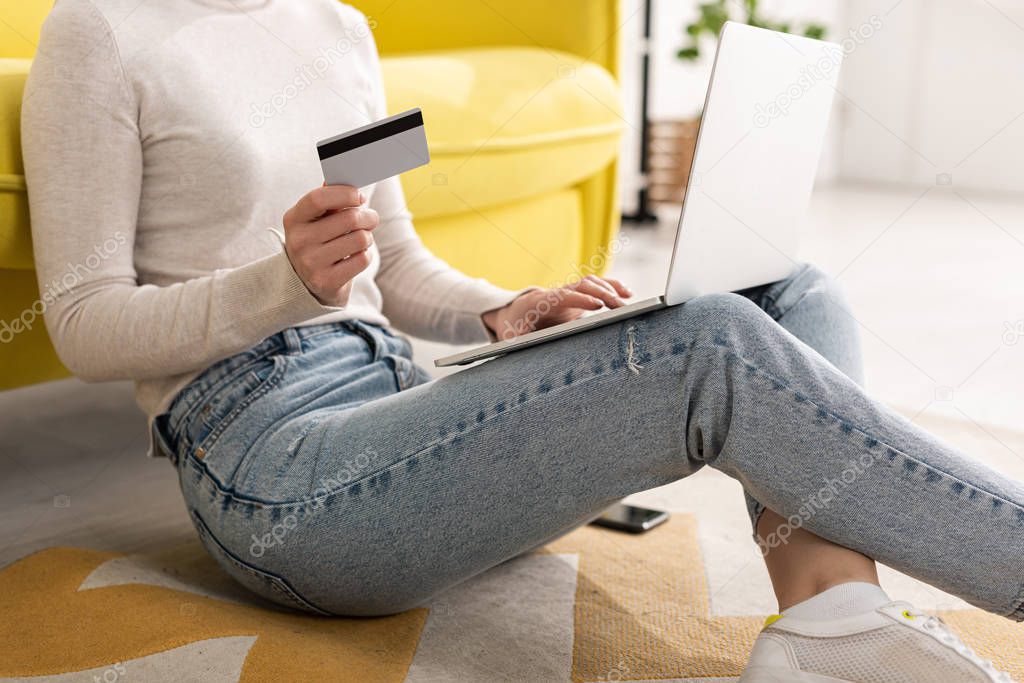 Cropped view of young woman holding credit card and using laptop on floor in living room