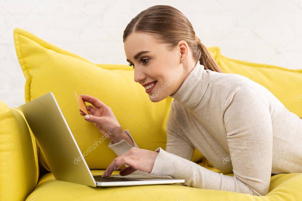 Side view of smiling woman using laptop and holding credit card while lying on couch 