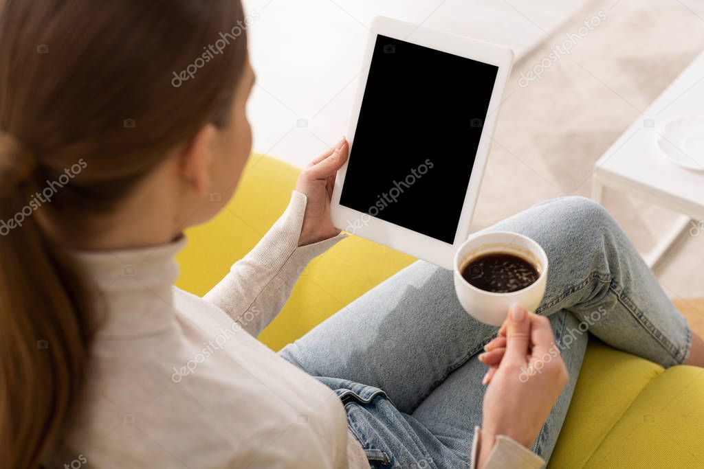 Over shoulder view of girl holding digital tablet with blank screen and drinking coffee on couch