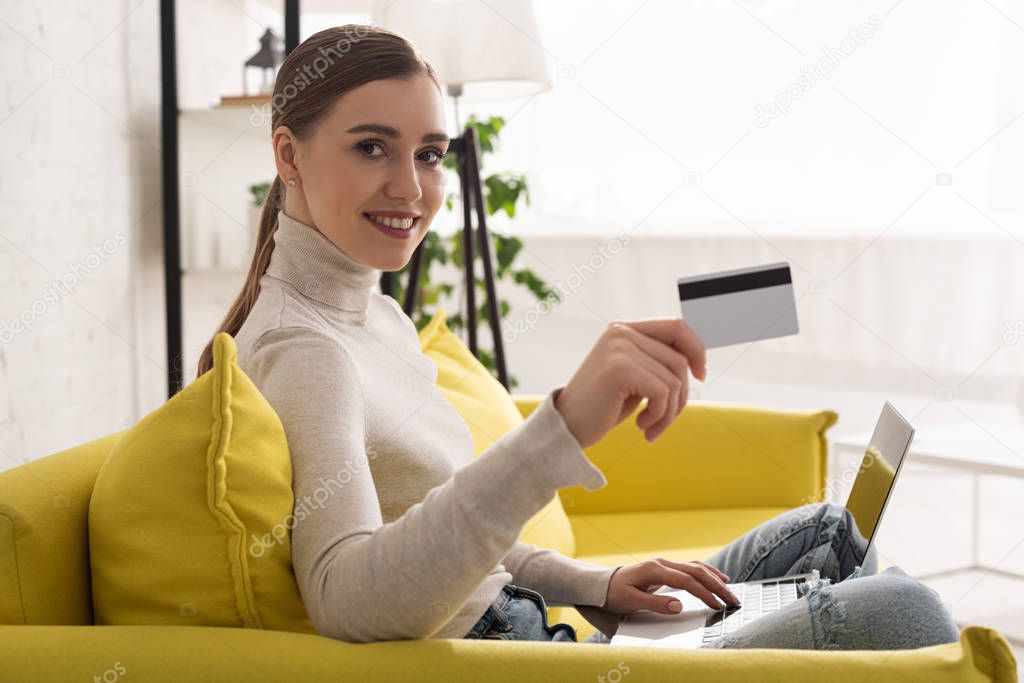 Smiling girl showing credit card and using laptop on couch in living room