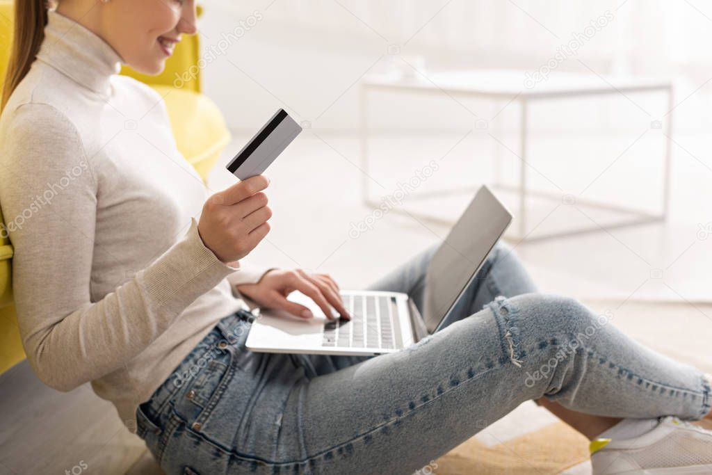 Cropped view of smiling woman holding credit card and using laptop on floor at home