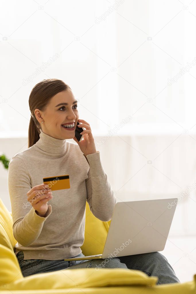 Smiling woman with credit card and laptop talking on smartphone on couch