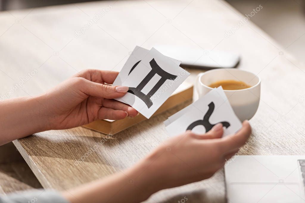 Cropped view of astrologer holding cards with zodiac signs beside coffee and book on table
