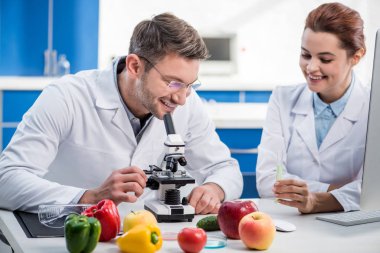 Gülümseyen moleküler beslenme uzmanı mikroskop kullanıyor ve meslektaşı test tüpü tutuyor. 