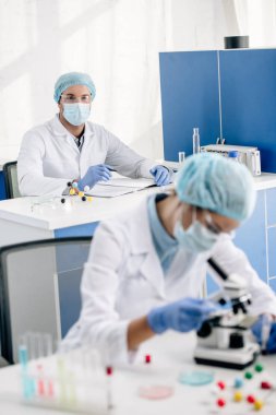 selective focus of genetic consultant looking at colleague with microscope clipart