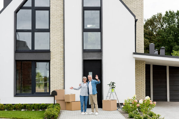 smiling mature man and woman waving near new house 
