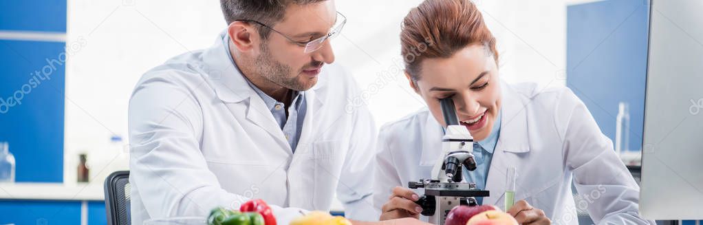panoramic shot of smiling molecular nutritionist using microscope and her colleague looking at her 