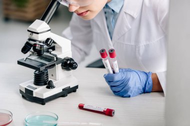 cropped view of genetic consultant using microscope and holding test tubes  clipart